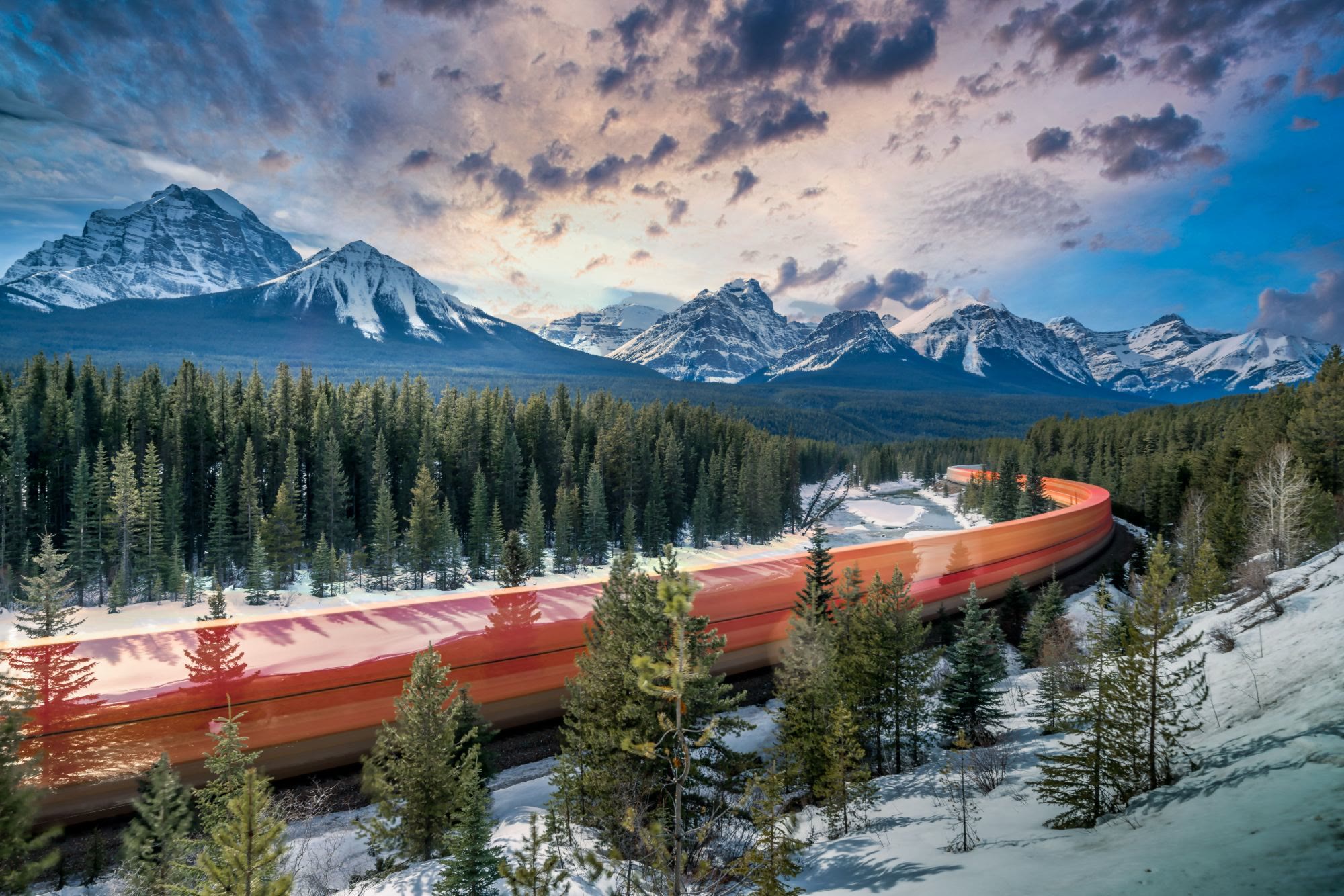 A train's light trail sweeps through snowy forest below the Canadian Rockies